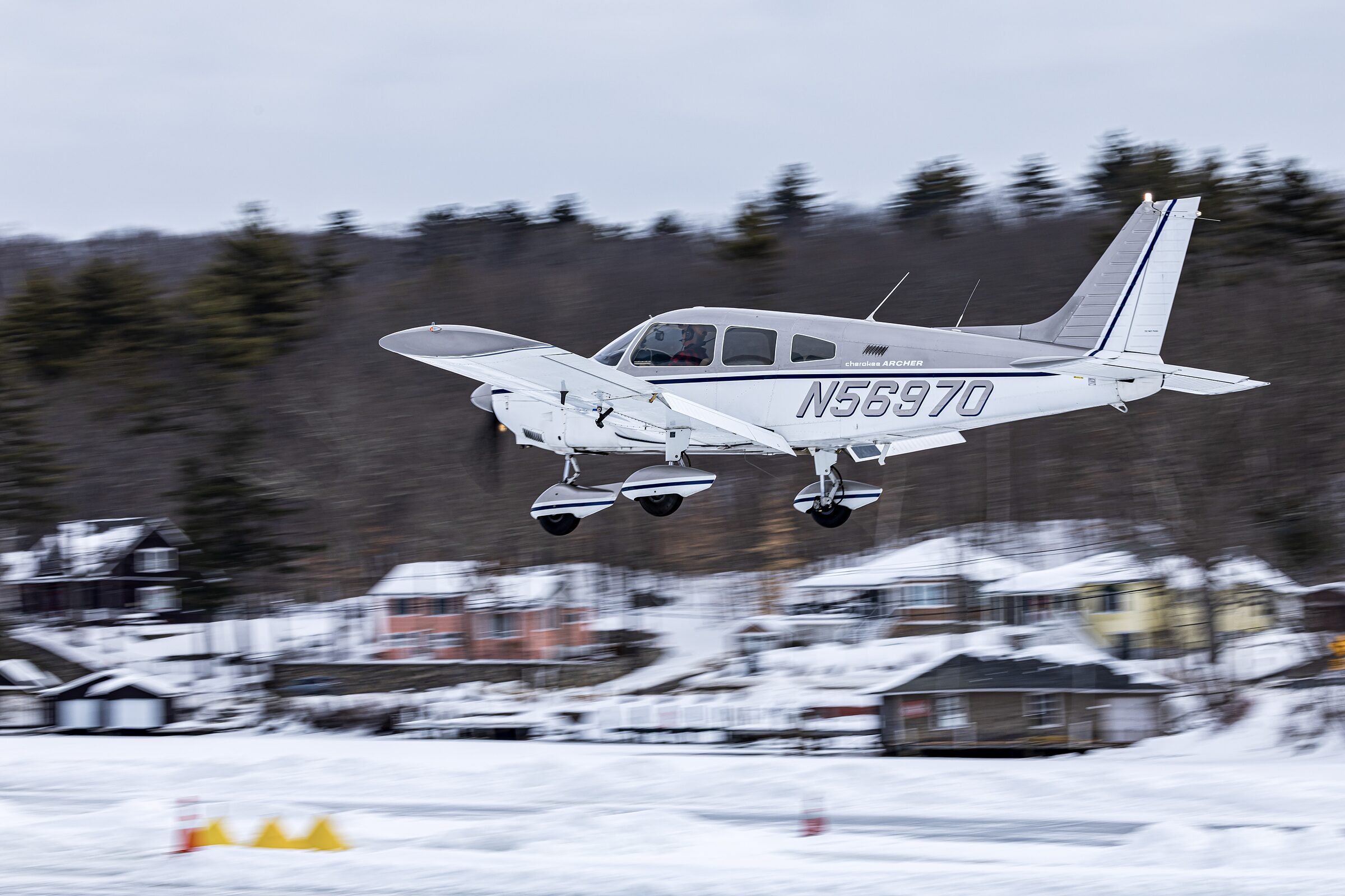 Elaine McPlane, a white Piper Cherokee Archer with a navy stripe, on short final over snow-covered lakefront houses at Alton Bay.